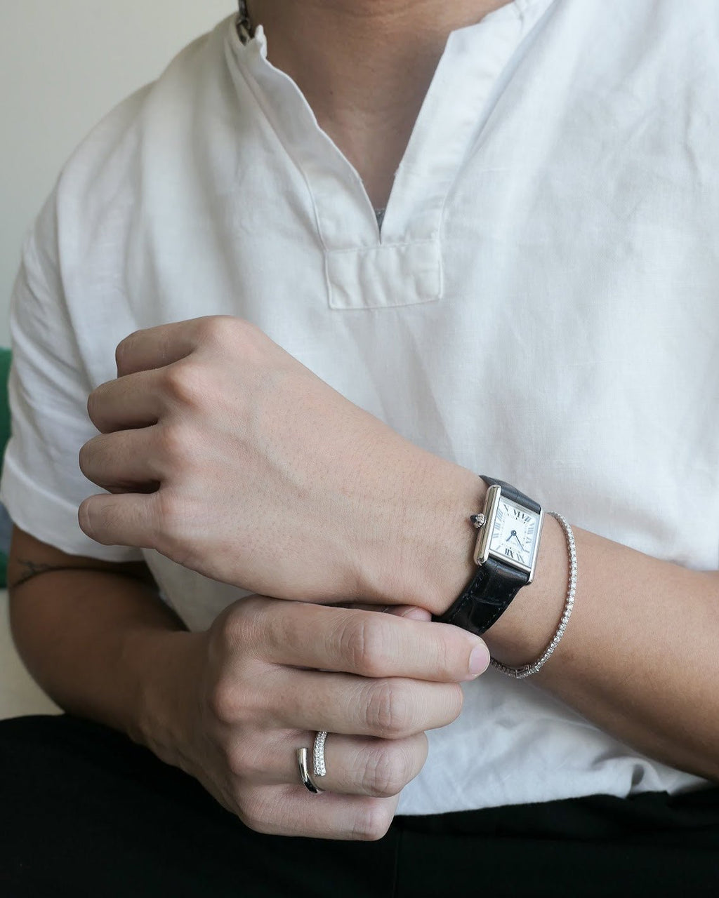 man neck down in a white linen shirt holding his hands stacked on each other, wearing a cartier watch and 18k white gold diamond tennis bracelet on one hand, and a diamond ring on the other hand