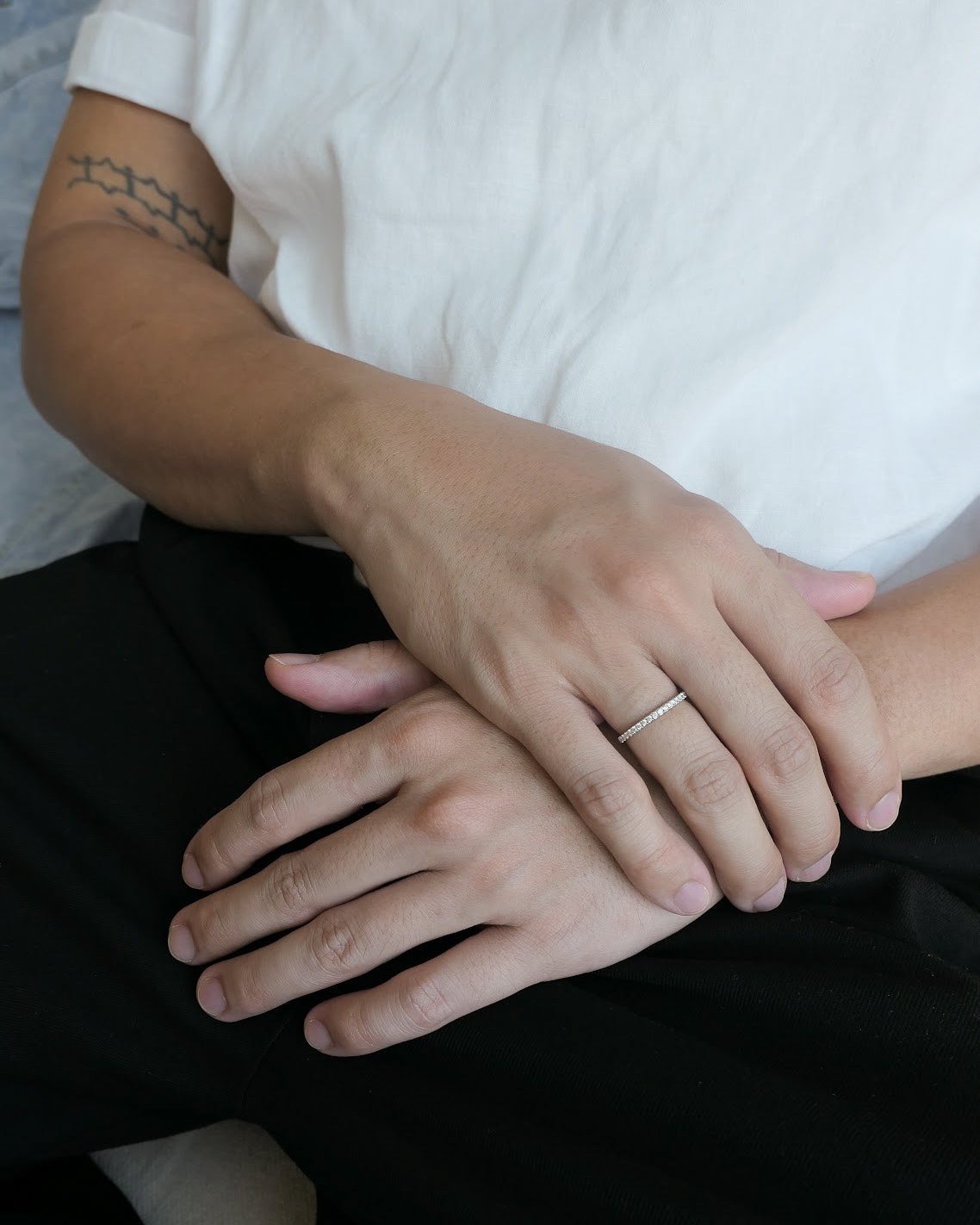 man in a white linen shirt waist down with a tattoo around his bicep, hands resting on each other with an 18k white gold diamond half eternity band ring on his finger