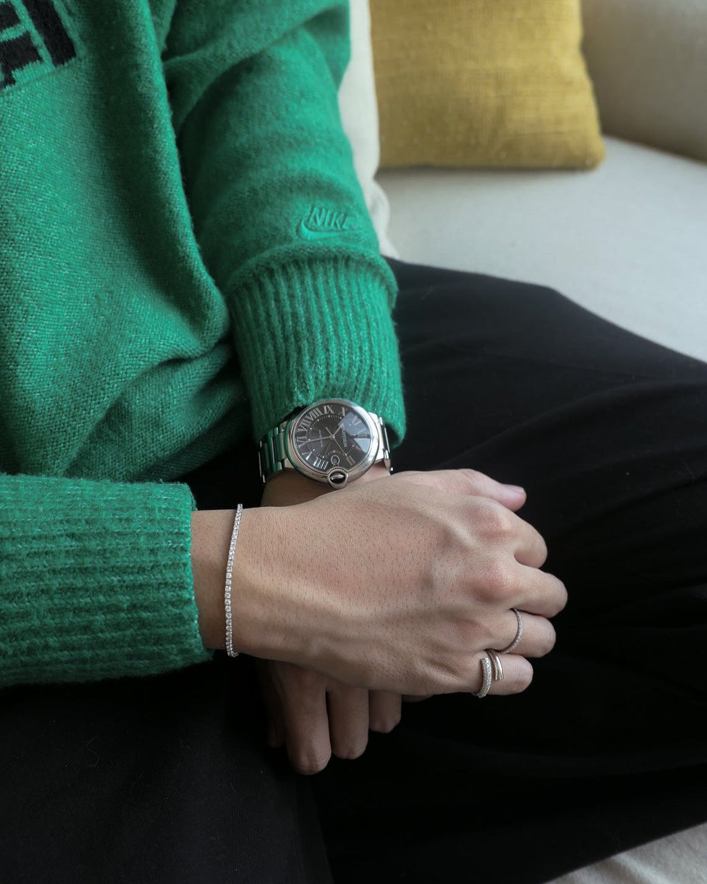 man sitting on couch waist down, wearing a green sweater and black pants, hand folded on his lap, wearing a cartier watch on one hand, wearing an 18k white gold diamond tennis bracelet and rings on the other hand