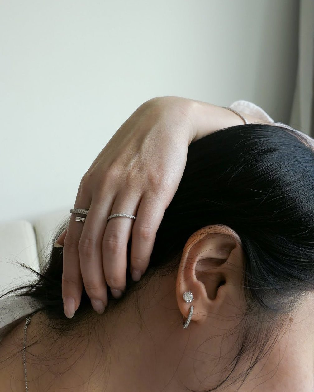 closeup of woman's ear, face down and hand draped over her black hair, ear has one 18k white gold diamond hoop and stud earrings on it, hand has two diamond rings on it