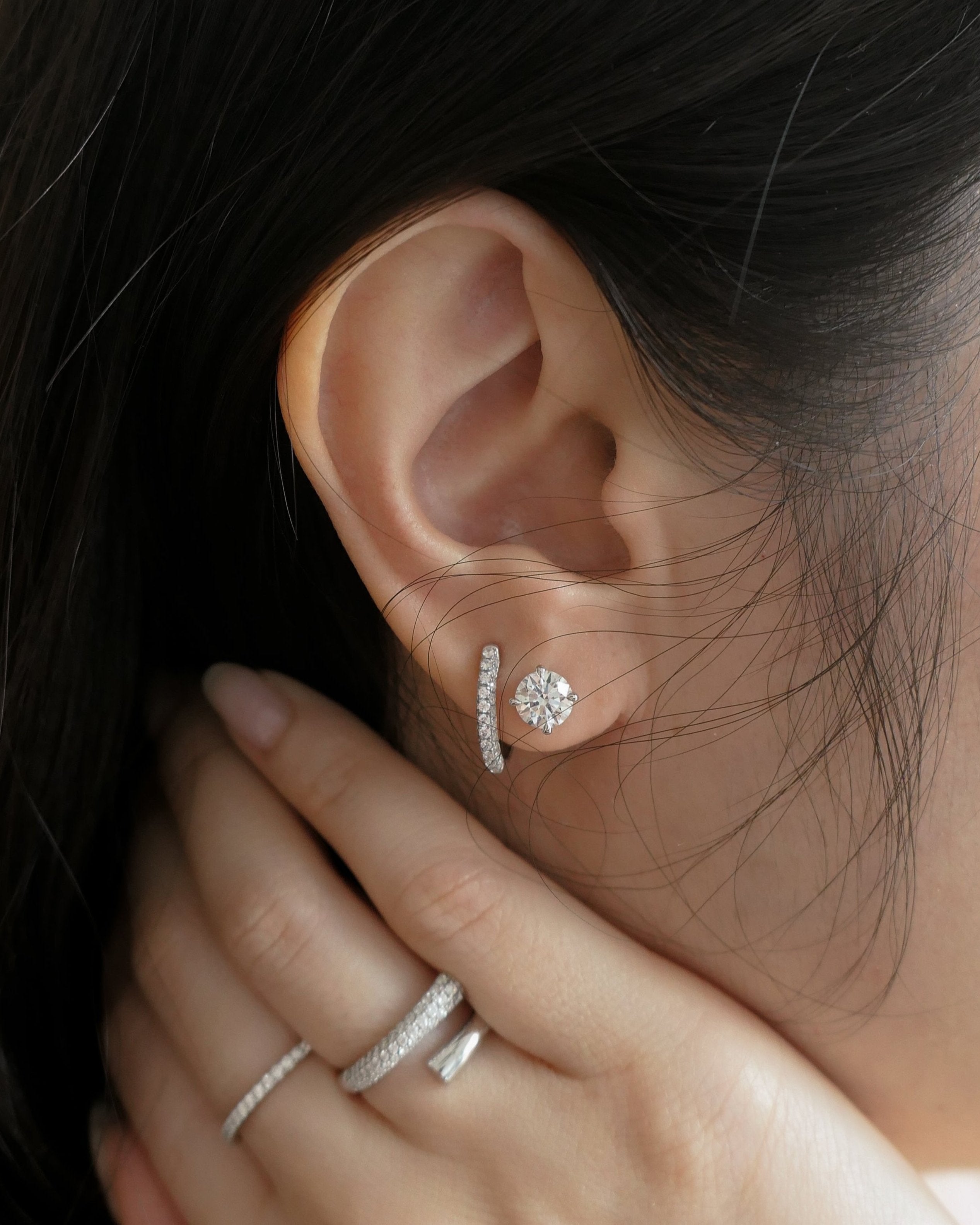 close up of woman's ear with hand on neck, ear has two diamond earrings on it, hand has two diamond rings on it