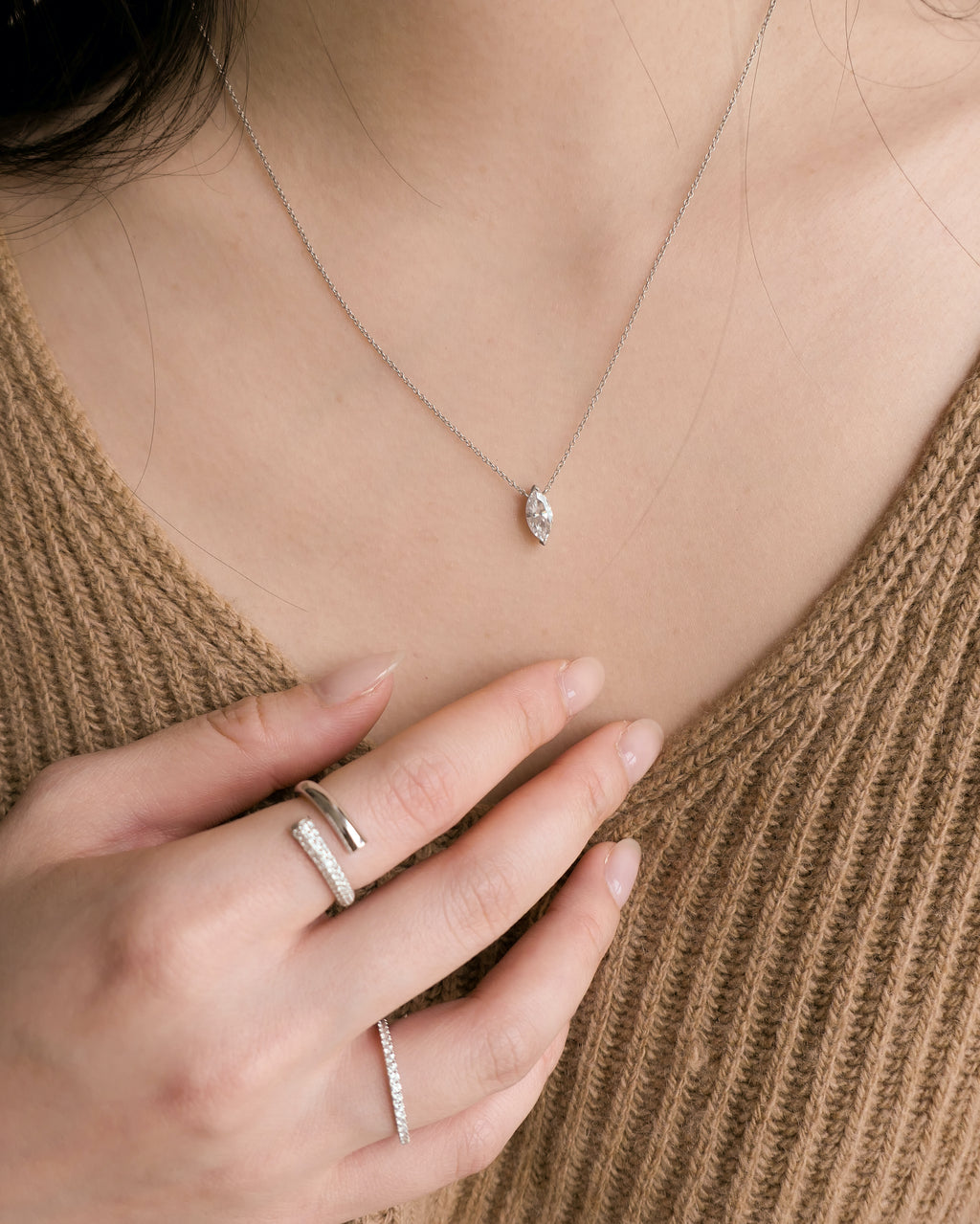 woman neck down in a brown cashmere sweater, hand held up to her chest, with two 18k white gold lab grown diamond rings on her fingers and a marquise diamond necklace on her neck